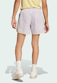 Helle lavendelfarbene Shorts mit elastischem Bund und gelbem Besatz. Verfügt über seitliche Streifen und eine Gesäßtasche. Getragen mit weißen Schuhen und Socken.