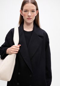 Abrigo negro de doble pecho con grandes solapas. El modelo sostiene un bolso tote de cuero crema. Textura suave, diseño estructurado y botones visibles.