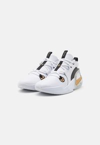 Baskets de sport blanches avec une tige texturée, des accents noirs, des éléments en or et une semelle rembourrée avec un logo Nike distinctif.