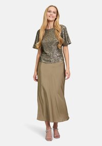 Błyszczący złoty top z krótkim rękawem ozdobiony cekinami, zestawiony z długą, beżową satynową spódnicą. Modelka ma na sobie przezroczyste sandały na obcasie. Gładkie tekstury i elegancki design.