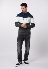 Hanorac cu glugă în nuanțe de bleumarin, alb și verde închis, cu finisaj texturat. Are un buzunar tip cangur și șnururi. Purtați cu blugi cu aspect uzat.