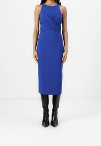 Blå ärmlös klänning med en vriden frontdesign, åtsittande silhuett, knä-lång, i kombination med svarta knähöga spetsiga stövlar.