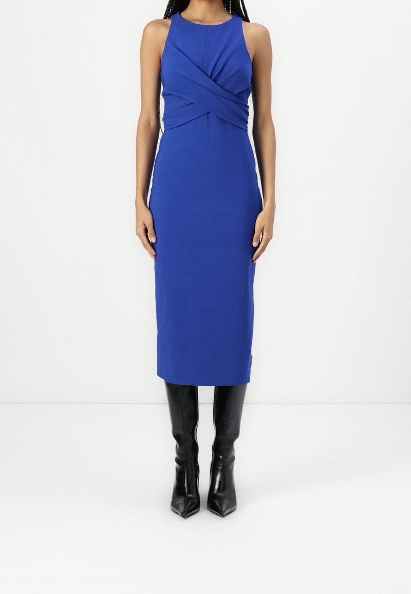 Blå ärmlös klänning med en vriden frontdesign, åtsittande silhuett, knä-lång, i kombination med svarta knähöga spetsiga stövlar.