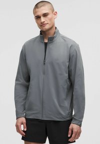 Veste grise avec col montant, fermeture éclair intégrale, poignets élastiques et tissu léger et lisse, associée à un short noir.
