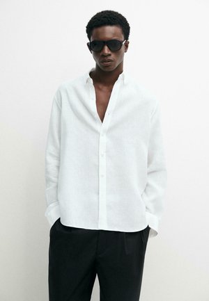 Giovane uomo che indossa occhiali da sole neri, una camicia bianca con bottoni parzialmente aperta e pantaloni neri, in piedi davanti a uno sfondo bianco semplice.