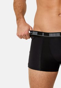 Man i svarta boxerkalsonger som drar i midjebandet bort från midjan, vilket visar passform och tygstruktur, överkropp och övre låren synliga.