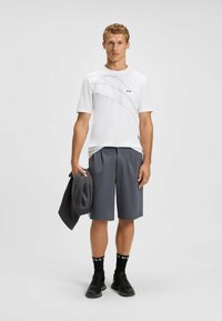 Hvid atletisk t-shirt med subtilt grå design og sort logo, parret med mørkegrå shorts. Modellen holder en mørk jakke og har sorte sko og sokker på.