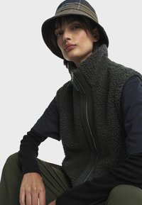 Gilet en polaire gris foncé avec col montant, fermeture éclair et surface texturée ; associé à une chemise à manches longues noire et un pantalon vert olive.