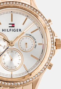 Reloj de oro rosa con esfera blanca, tres subesferas y un bisel decorado con cristales. El logotipo de la marca se muestra de forma destacada en el centro.