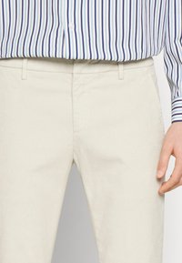 Světlé béžové bavlněné kalhoty s žebrovanou strukturou, se standardním pasem a bočními kapsami. V kombinaci s modře pruhovanou košilí na zapínání na knoflíky.