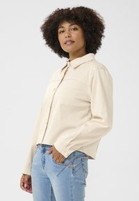 Beige kortärmad knäppskjorta med krage, långa ärmar och en slät yta, kombinerad med ljusblå jeans med sneda sömmar.