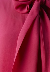 Rosa chiffonstof med en glat tekstur, blødt draperede folder og en sløjfedetail ved halsudskæringen. Farven er livlig og klar.