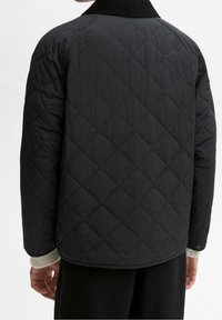 Musta tikattu takki, jossa on ribbineulottu kaulus, teksturoitu kuvio, napsautettavat kiinnikkeet ja rento istuvuus. Ranteessa näkyvä neuleen hihansuu.