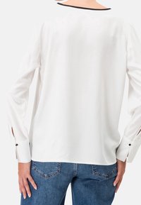 Chemisier blanc à manches longues avec une coupe décontractée, comportant des bordures noires au niveau de l'encolure et des poignets à boutons, associé à un jean en denim bleu.