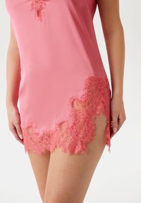 Rosa satin-nattklänning med detaljerad spetskant vid nederkanten, med en slät textur och en avslappnad passform. Spetsdetaljerna skapar en öppen design.