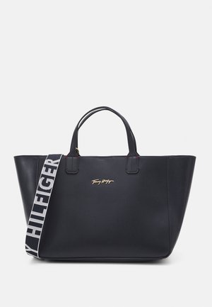 Sort tote taske lavet af kunstlæder med to øverste håndtag, en aftagelig rem med "Tommy Hilfiger"-logo og subtile røde syninger som detaljer.