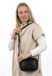 Borsa a tracolla in pelle nera con dettagli intrecciati, forma rettangolare, chiusura con zip e tracolla regolabile. Il modello indossa un top beige testurizzato.