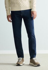 Sötétkék farmernadrág, slim fit szabással, bézs sportcipőkkel, fehér cipőfűzőkkel és texturált talppal párosítva. Kábeles kötésű krémszínű pulóver.