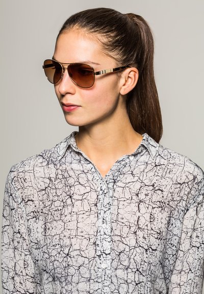 Brown, gradient sunglasses with a slim gold frame. Worn with a patterned white and black shirt featuring a crackle design.