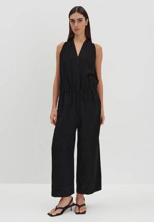 Black sleeveless jumpsuit with a V-neckline, cinched waist, wide legs, and smooth fabric. Paired with black strappy sandals.