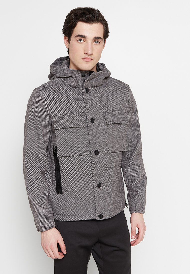Grijze, textuurjacket met een capuchon, voorzien van twee voorzakken en een met rits gesloten zijzak, dichtgemaakt met zwarte knopen.