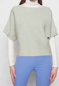 Ljusgrön, texturerad stickad topp med korta, breda ärmar. Bärs över en vit polotröja, ihop med blå byxor. Enkel, modern design.