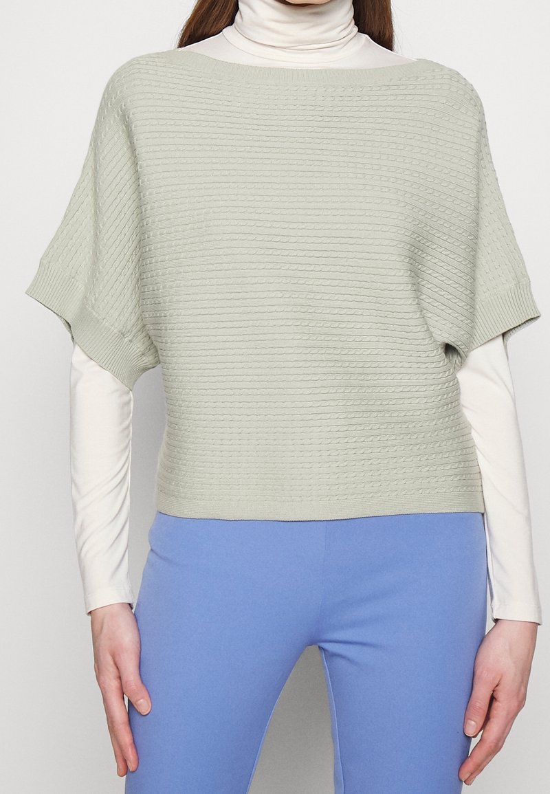 Ljusgrön, texturerad stickad topp med korta, breda ärmar. Bärs över en vit polotröja, ihop med blå byxor. Enkel, modern design.