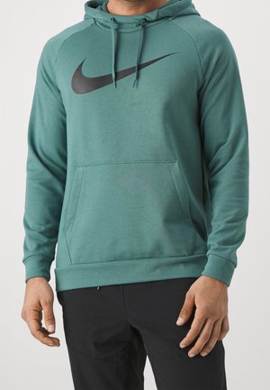 Man draagt een groenblauwe Nike-hoodie met zwart swoosh-logo, gecombineerd met zwarte broek, staand tegen een effen achtergrond.