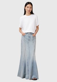 Maxi gonna in denim azzurro chiaro con dettagli a cucitura verticale, abbinata a una t-shirt bianca a maniche corte. Il modello posa con le mani in tasca.