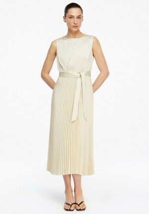 Kobieta z włosami związanymi do tyłu, ubrana w kremową plisowaną sukienkę midi bez rękawów z paskiem w talii, stojąca z rękami za plecami i czarnymi sandałami.