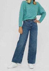 Pull turquoise côtelé associé à un jean en denim bleu à taille haute et jambes larges, accompagné de baskets blanches. Design simple avec une coupe décontractée et sans motifs.