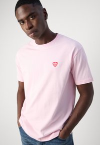 Roze katoenen t-shirt met een klein rood hartlogo en de tekst "MAKIA". Korte mouwen en een ronde hals, casual pasvorm.