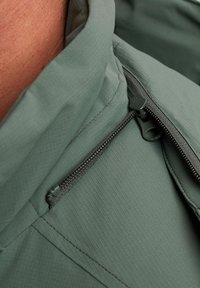 Giacca verde leggera con tessuto testurizzato, dotata di chiusura con zip e colletto alto. Includo un primo piano del dettaglio della zip.