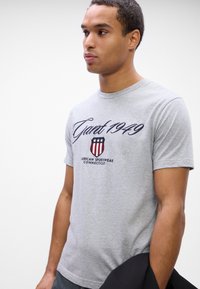 T-shirt de algodão cinza com a inscrição "Grant 1949" em letras cursivas e um emblema com listras vermelhas e azuis, mangas curtas e decote redondo.