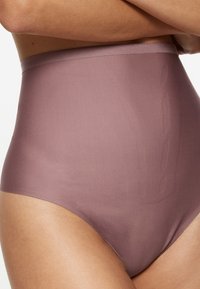 Vrouw draagt hooggetailleerde, naadloze mauve corrigerende slip, afgebeeld van torso tot bovenbenen tegen een witte achtergrond.