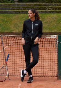 Musta urheiluasu, jossa on vetoketjullinen takki ja logo, slim-fit-housut, yhdistettynä mustiin lenkkareihin. Tennis Maila ja verkko taustalla.
