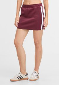 Gonna sportiva bordeaux con vita elástica, strisce bianche ai lati e texture morbida, indossata con scarpe da ginnastica chiare con strisce nere.