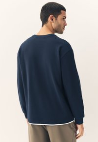 Navy long-sleeve sweatshirt with a textured ribbed surface, round neckline, and contrasting white hem, paired with light-colored trousers.