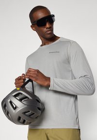 Light gray long-sleeve shirt with "ENDURA" logo, holding a dark gray helmet with ventilation holes and yellow accent. Black sunglasses.