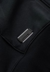 Gros plan sur un tissu noir avec une étiquette métallique rectangulaire noire et argentée marquée « ombre » cousue sur la couture.