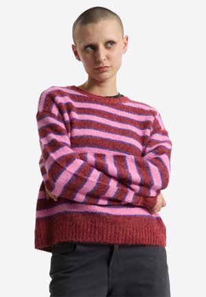 Person med barberet hoved iført en lyserød og rød striped fuzzy sweater, korslagte arme, som kigger til siden med et neutralt udtryk.