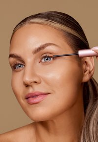 Applicatore di mascara con un pennello sottile in uso sulle ciglia. Il contenitore è rosa e lo sfondo ha una tonalità beige calda. Texture della pelle liscia.