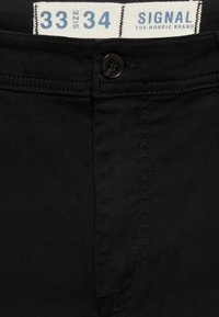 Zwarte jeans met een gladde, matte textuur. Voorzien van een knoop en stiksel detail. Label toont maat 33/34 van het merk "Signal."