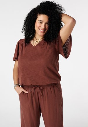 Femme aux cheveux bouclés souriante, portant un haut marron à manches courtes et un pantalon assorti, une main dans la poche et l'autre touchant sa tête.