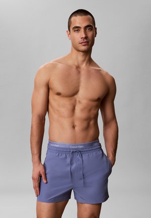 Muskelöser Mann mit kurzen Haaren, der blaue Calvin Klein Badeshorts trägt und mit einer Hand in der Tasche vor einem neutralen Hintergrund steht.