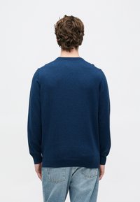 Σκούρο μπλε πουλόβερ με μακριά μανίκια, στρογγυλή λαιμόκοψη και μαλακή υφή. Η πίσω όψη δείχνει την εφαρμοστή μέση και τα ριμπ μανικία. Φοριέται με ανοιχτό μπλε τζιν.