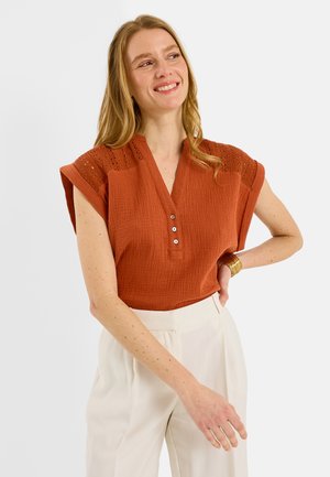 Mujer sonriente, vestida con una blusa con textura de color óxido-anaranjado con hombros de encaje y pantalones blancos de talle alto, posando con la mano en la cadera.