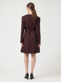 Γυναίκα με τα μαλλιά της δεμένα πίσω, φοράει ένα σκουρόχρωμο καφέ παλτό μήκους μέχρι τα γόνατα με ζώνη και μαύρα παπούτσια με τακούνια, στέκεται γυρισμένη προς τα πίσω σε απλό φόντο.