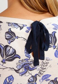 Nahaufnahme eines Oberteils mit floralem Muster in Blau und Schwarz, das eine marineblaue Stoffschleife zeigt, die am hinteren Ausschnitt auf der Schulter einer Person gebunden ist.