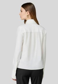 Camicetta bianca a maniche lunghe con un sottile lucido, caratterizzata da una cucitura a yoke posteriore, polsini con bottone e una vestibilità sartoriale, abbinata a pantaloni neri.